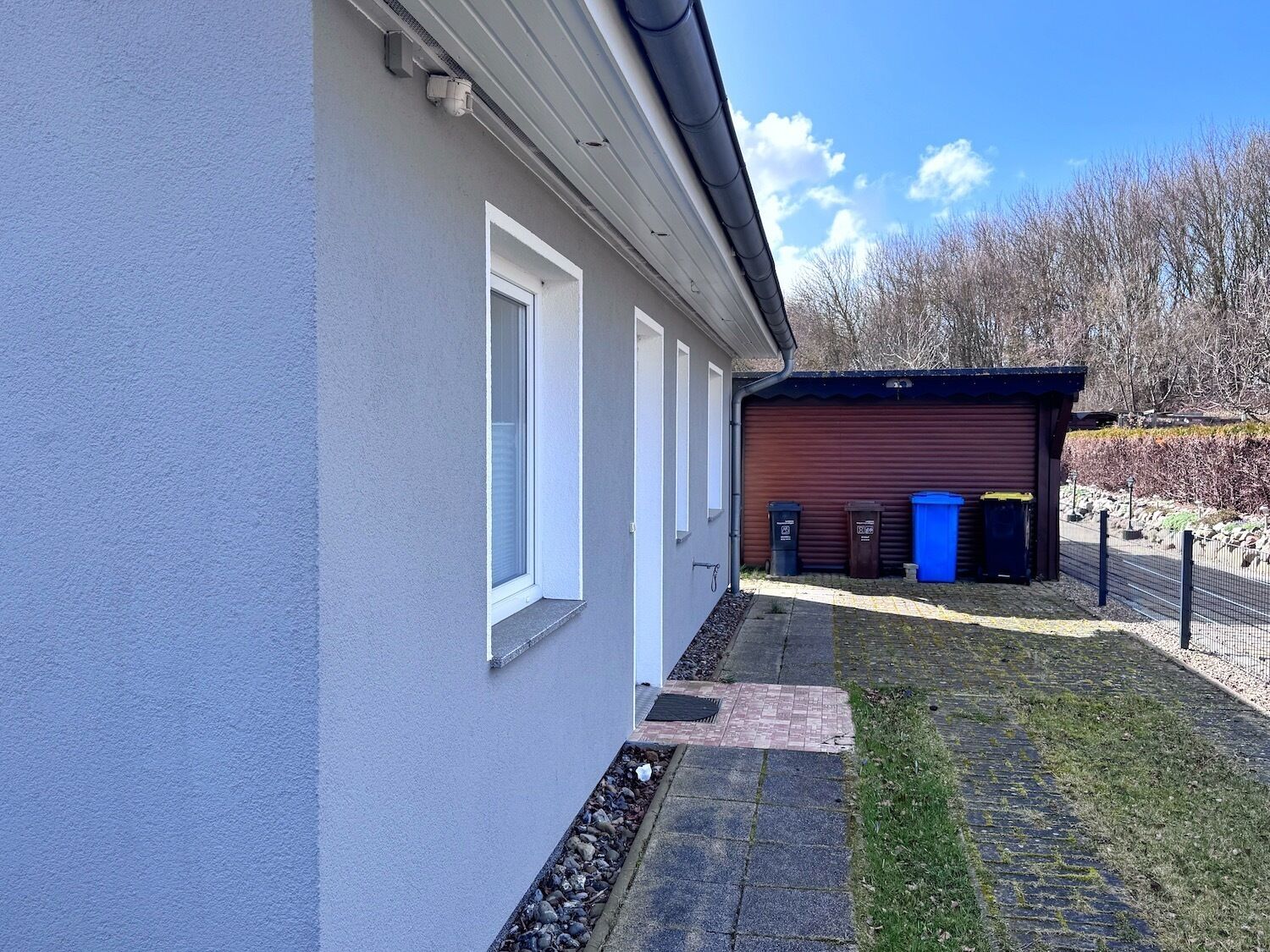 Um edifício com fachada cinzenta, uma porta de garagem e uma pequena área coberta com lixeiras.