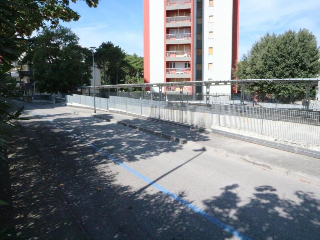Un edificio de varios pisos con balcones, un área pavimentada con una cerca y árboles proyectando sombras.