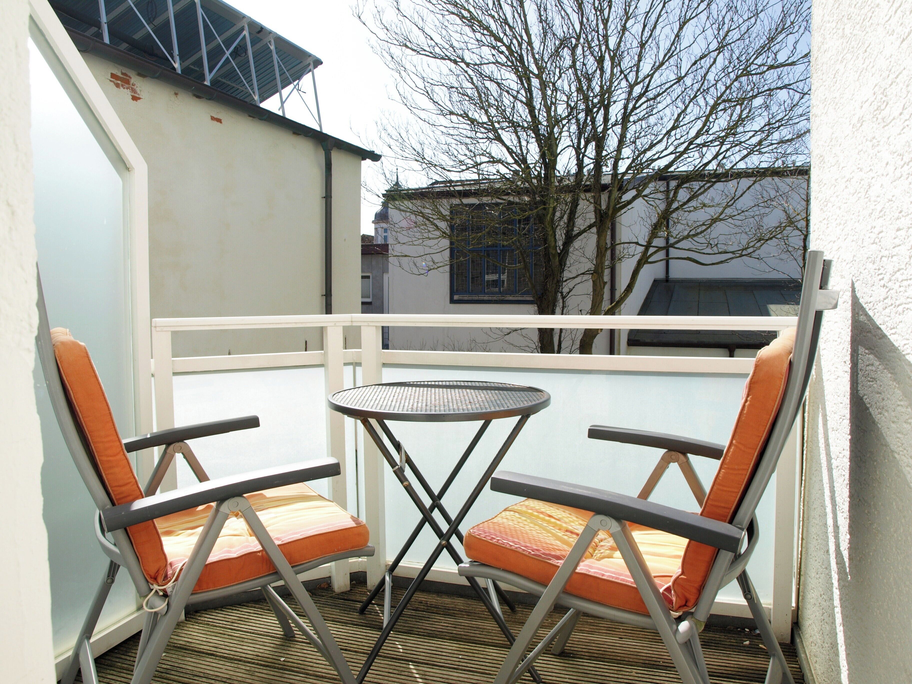Ein Balkon mit zwei Stühlen und einem kleinen Tisch, mit Blick auf ein Gebäude und einen Baum.