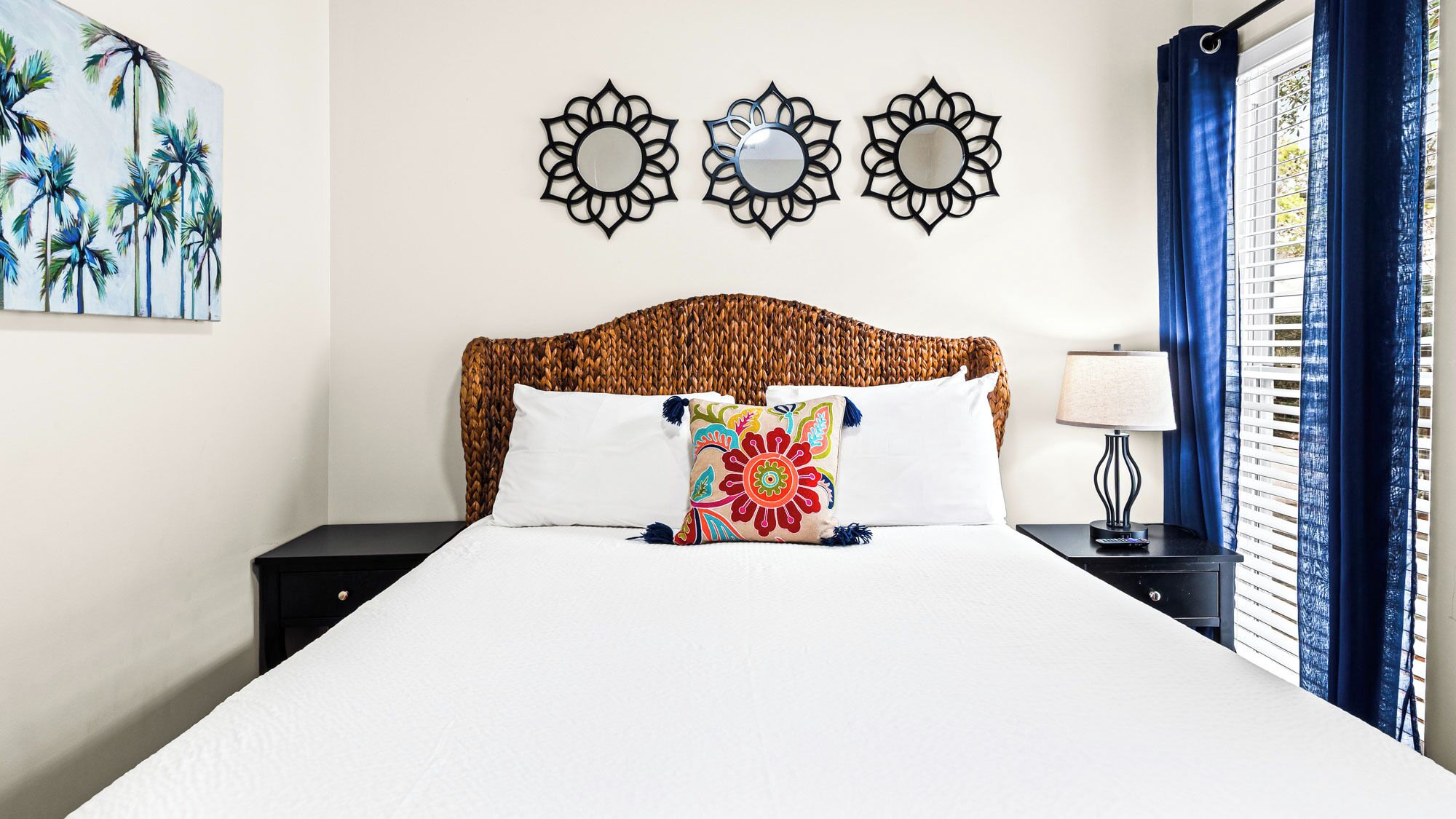 Ein Hotelzimmer mit einem gewebten Kopfteil, einem Bett mit weißen Leinentüchern, dekorativen Kissen, einem Nachttisch mit einer Lampe und Wandbildern, darunter ein Palmenbaumgemälde und drei sonnenförmige Spiegel.