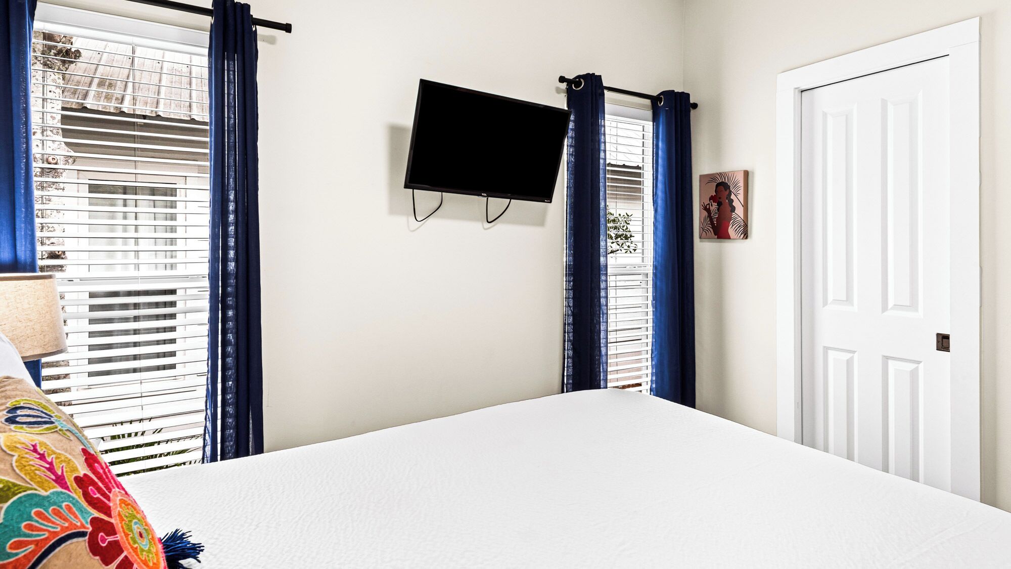 A bedroom with a bed, a TV mounted on the wall, a door, and blue curtains.