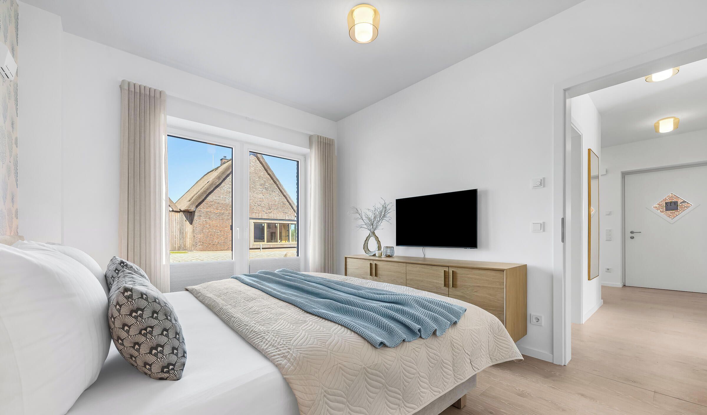 A bedroom with a bed, a TV, a wooden dresser, and a view of a brick building outside.