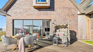 Een bakstenen huis met een houten terras, buiten zitplaatsen en een schuifdeur naar een binnenruimte.