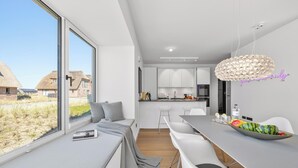 Een moderne keuken met witte kasten, een eethoek met een groot raam en uitzicht op huizen met rieten daken.