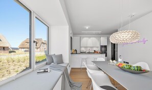 A modern kitchen with white cabinets, a dining area with a large window, and a view of thatched-roof buildings outside. - Premium vacation home Halber Deich in the dune park with terrace and sauna (List auf Sylt)