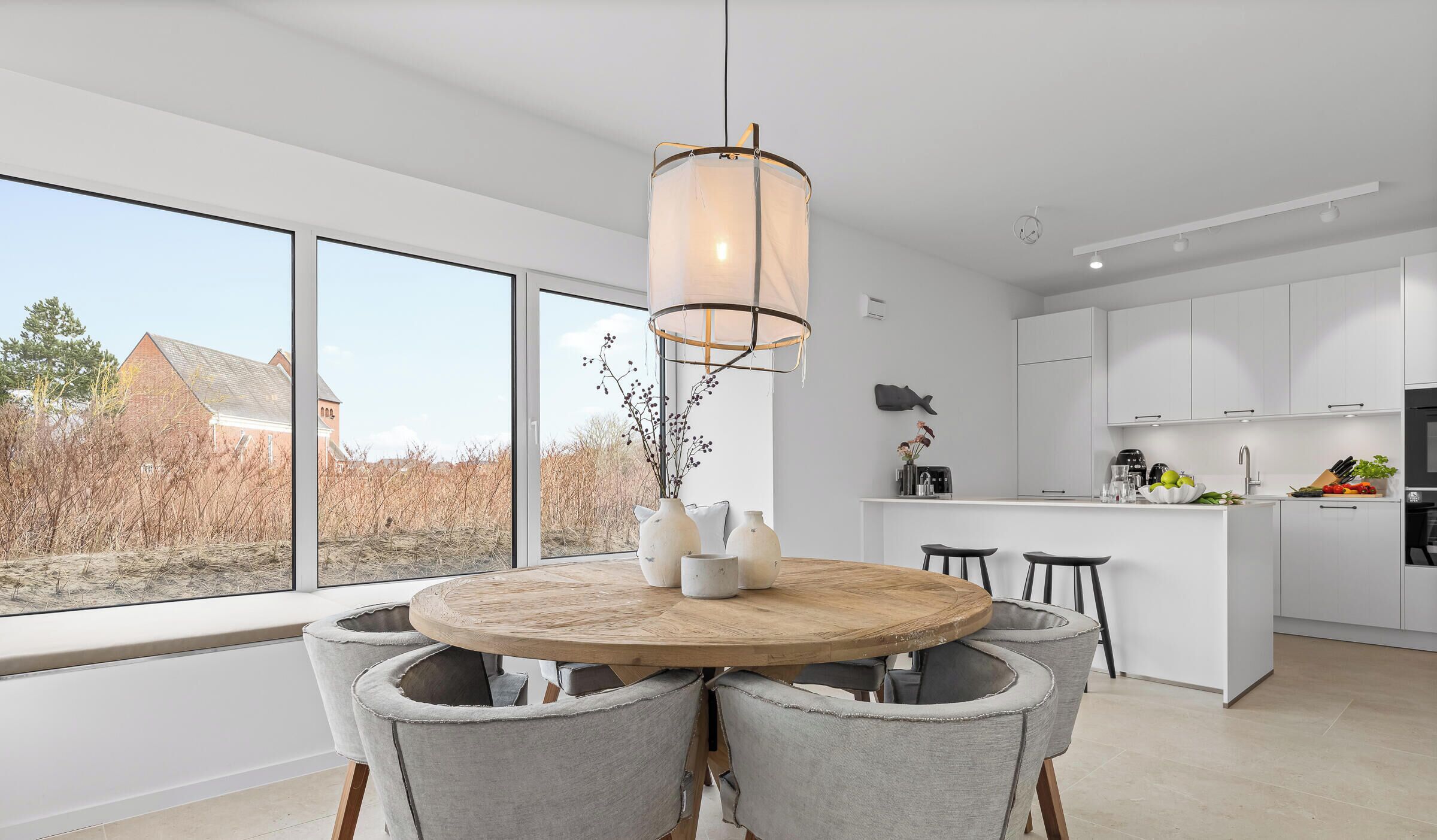 Een moderne keuken met witte kastjes, een houten eettafel met vier stoelen, en een groot raam met uitzicht op een gebouw en bomen.