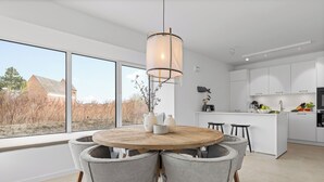 A modern kitchen with white cabinets, a wooden dining table with four chairs, and a large window offering a view of a building and trees. - Premium vacation home Halber Deich in the dune park with terrace and sauna (List auf Sylt)