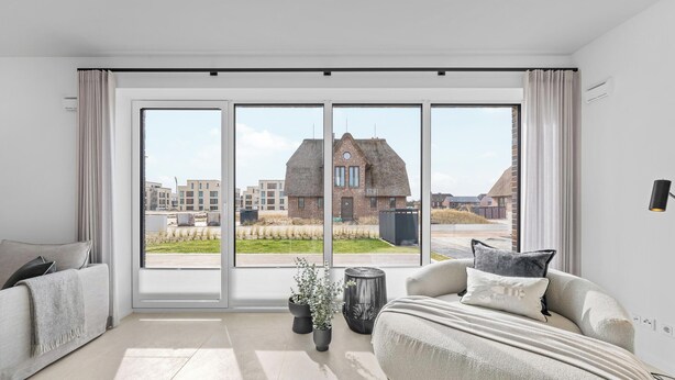 A modern living room with a large sliding glass door, a grey sofa, a black side table, and a potted plant. - Premium vacation home Halber Deich in the dune park with terrace and sauna (List auf Sylt)