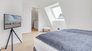 A bedroom with a bed, a television on a tripod, a window, and a door leading to a bathroom. - Premium vacation home Haus Deich in the dune park with terrace and sauna (List auf Sylt)