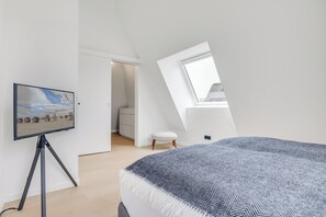 A bedroom with a bed, a television on a tripod, a window, and a door leading to a bathroom. - Premium vacation home Haus Deich in the dune park with terrace and sauna (List auf Sylt)