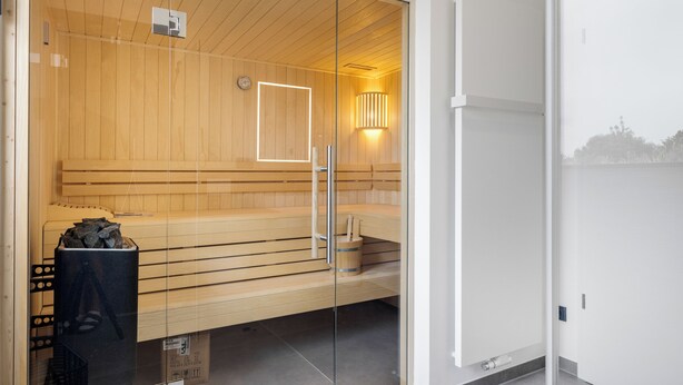 A sauna with wooden benches, a glass door, and a wall-mounted light fixture. - Premium vacation home Haus Deich in the dune park with terrace and sauna (List auf Sylt)