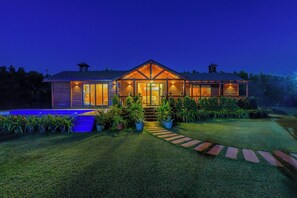 A two-story house with a well-maintained lawn, a pathway leading to the entrance, and a pool on the left. - ELIVAAS Riveira Hills - 3 BHK Cottage (Lachhmangarh)