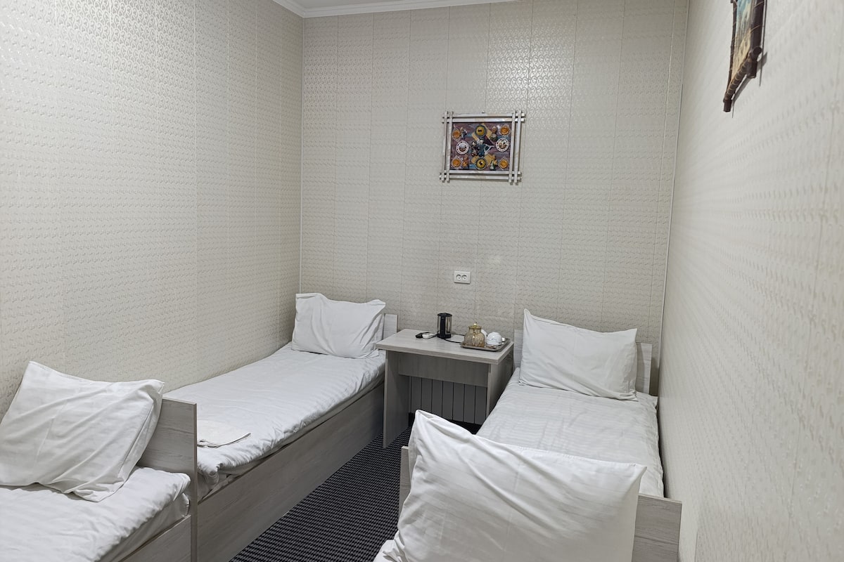 A small, single-room hotel with two single beds, a small table with a lamp, and a decorative item on the wall.