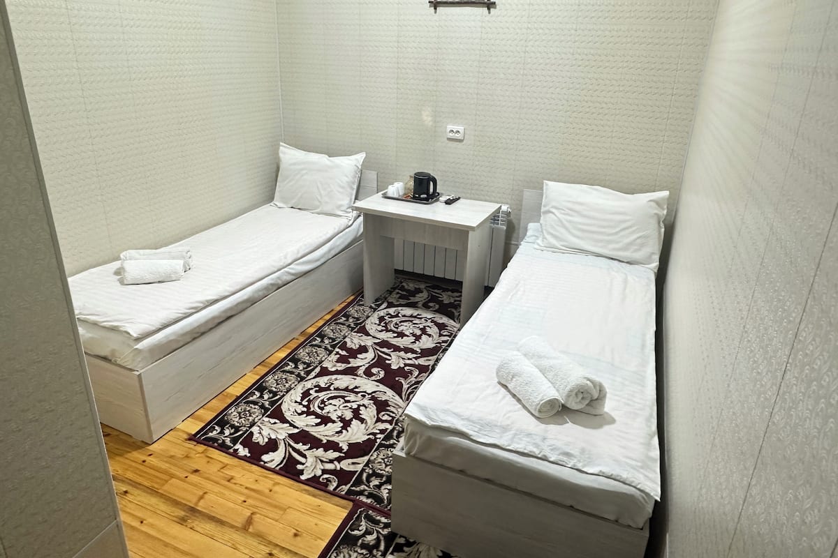 A small, clean room with two single beds, a small table with a kettle, and a patterned rug.