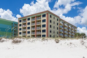 Un hôtel à plusieurs étages, doté de balcons à chaque niveau, situé sur une plage de sable fin, sous un ciel dégagé.