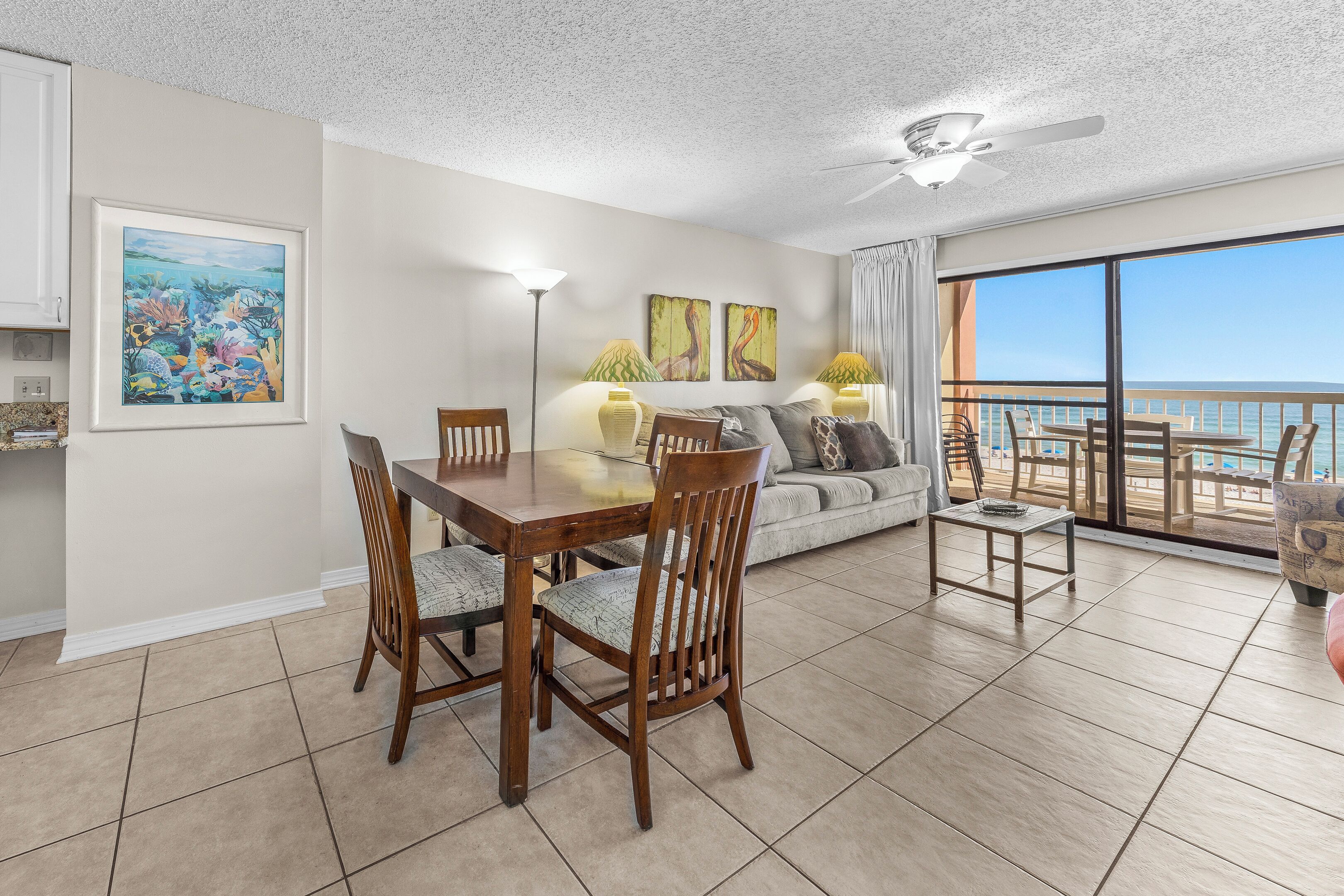 A dining area with a table and chairs, a sofa, a ceiling fan, and a view of the ocean through large windows.