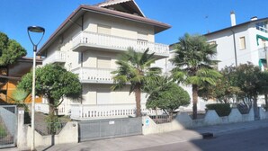 Un edificio residencial de varios pisos con balcón, palmeras y entrada protegida por un portón.