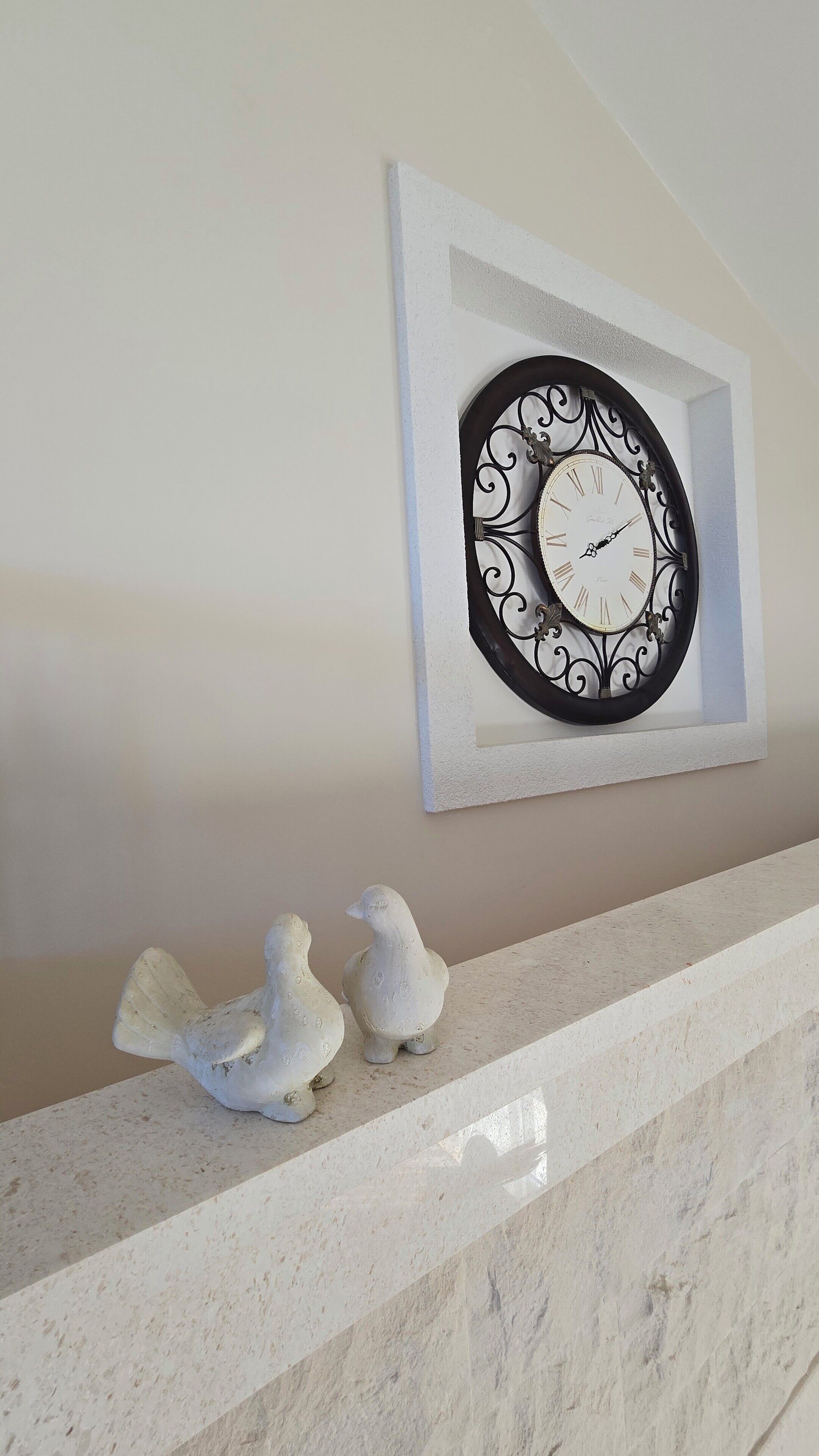 A wall-mounted clock with a decorative frame, two white bird figurines on a marble ledge, and a plain wall.