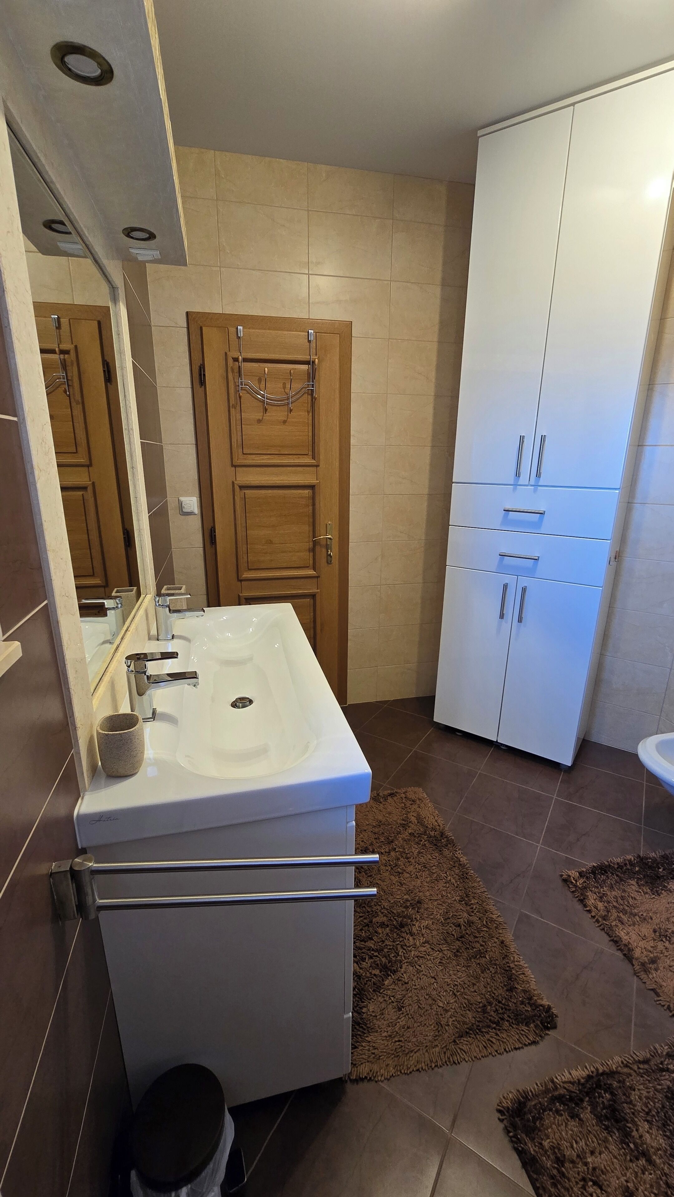 A bathroom with a white sink, a mirror, a wooden door, and a white wardrobe.