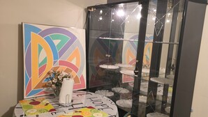 Ein Essbereich mit einem runden Tisch, der mit einer weißen Tischdecke und schwarzem Text bedeckt ist, einem schwarzen Schrank mit Glastüren und einem farbenfrohen, abstrakten Gemälde an der Wand.