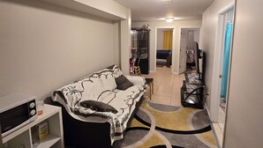 Ein Wohnzimmer mit einer Couch, einem kleinen Tisch, einer Mikrowelle und einem Regal. Ein Flur führt zu einem weiteren Zimmer.