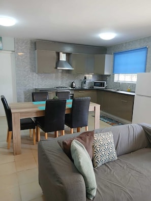 A modern kitchen with a dining area, a sofa with patterned cushions, and a window with a blue blind. - Casa Carmelita Maontanja Rey (Willemstad)