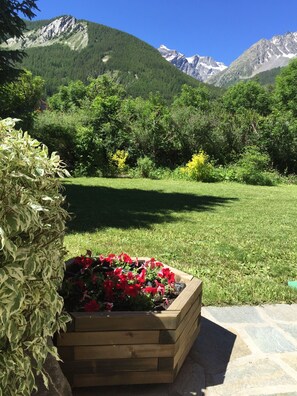 A garden with a wooden planter bed containing red flowers, surrounded by green grass and trees, with a mountainous backdrop. - Garden-level chalet at the foot of the ski slopes (Le Monêtier-les-Bains)