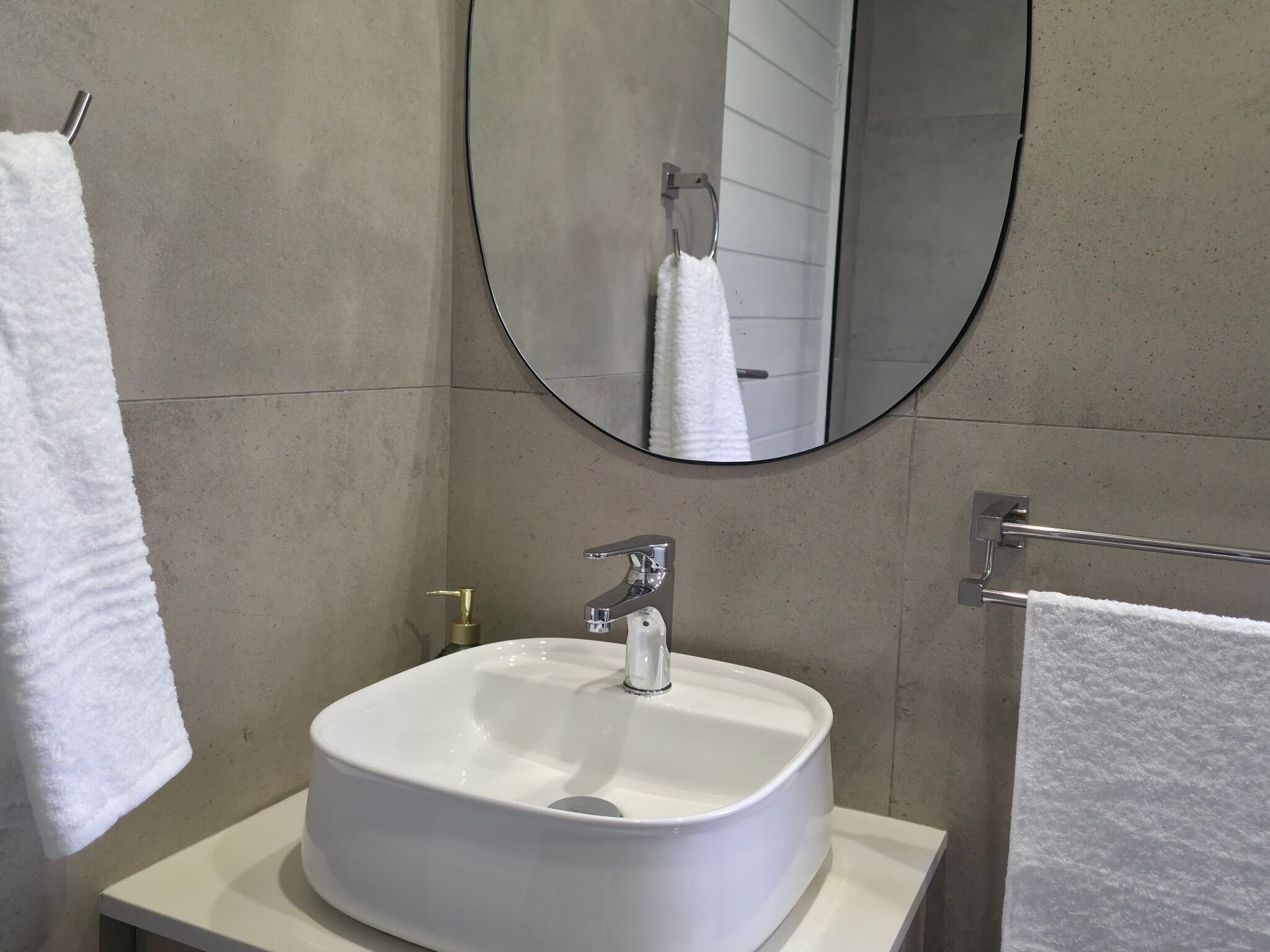 Un baño con un lavamanos blanco, un espejo redondo y dos toallas en percheros.