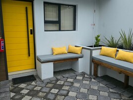 Un rincón con asientos tipo banca y cojines, una ventana con persianas y un extintor de incendios sujeto a la pared.