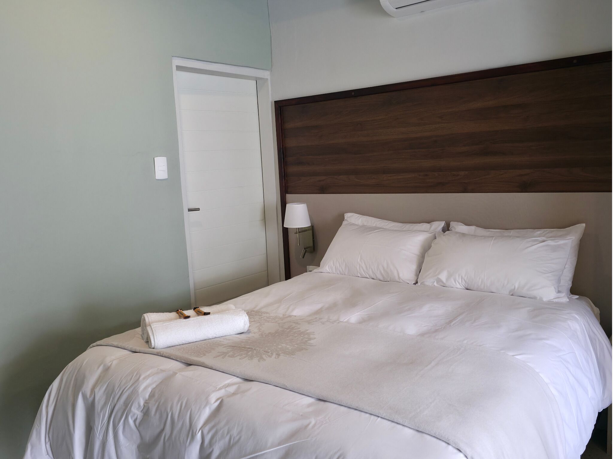 A neatly made bed with white linens, a wooden headboard, a bedside lamp, and a door with a light switch.