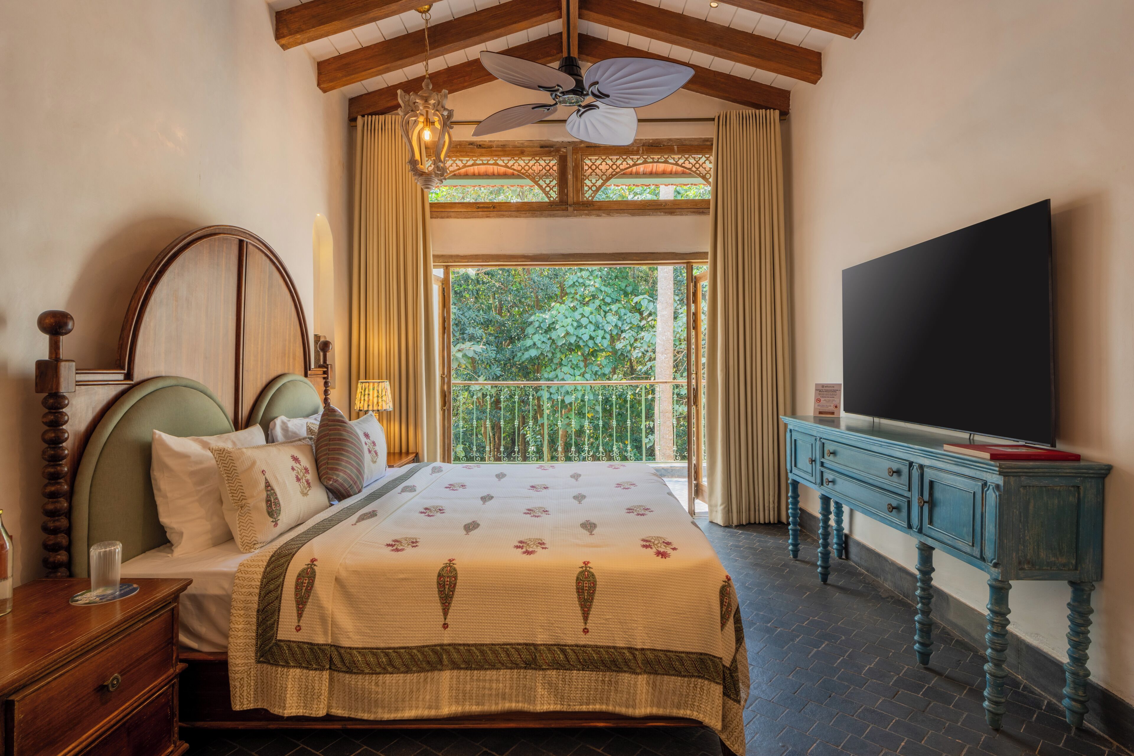 Une chambre avec un lit, une télévision, un balcon donnant sur la verdure et un ventilateur de plafond.