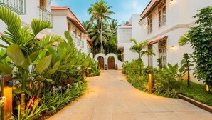 A pathway leading to a building with white walls and a red-tiled roof, surrounded by lush greenery and palm trees.