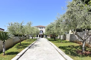 Un chemin de pierre mène à un bâtiment dont le toit est recouvert de tuiles, entouré d’arbres et d’un mur de pierre.