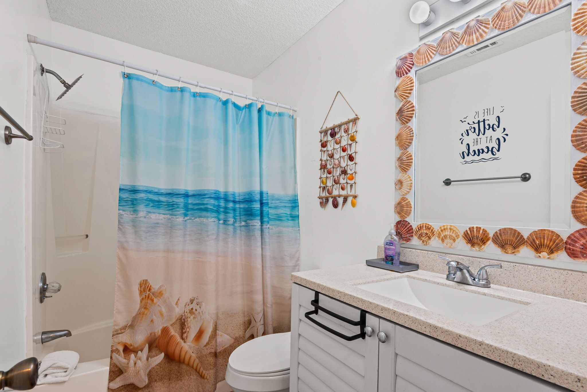 Un baño con cortina de ducha que muestra olas del océano, un inodoro, un lavabo con espejo y un adorno de conchas marinas en la pared.