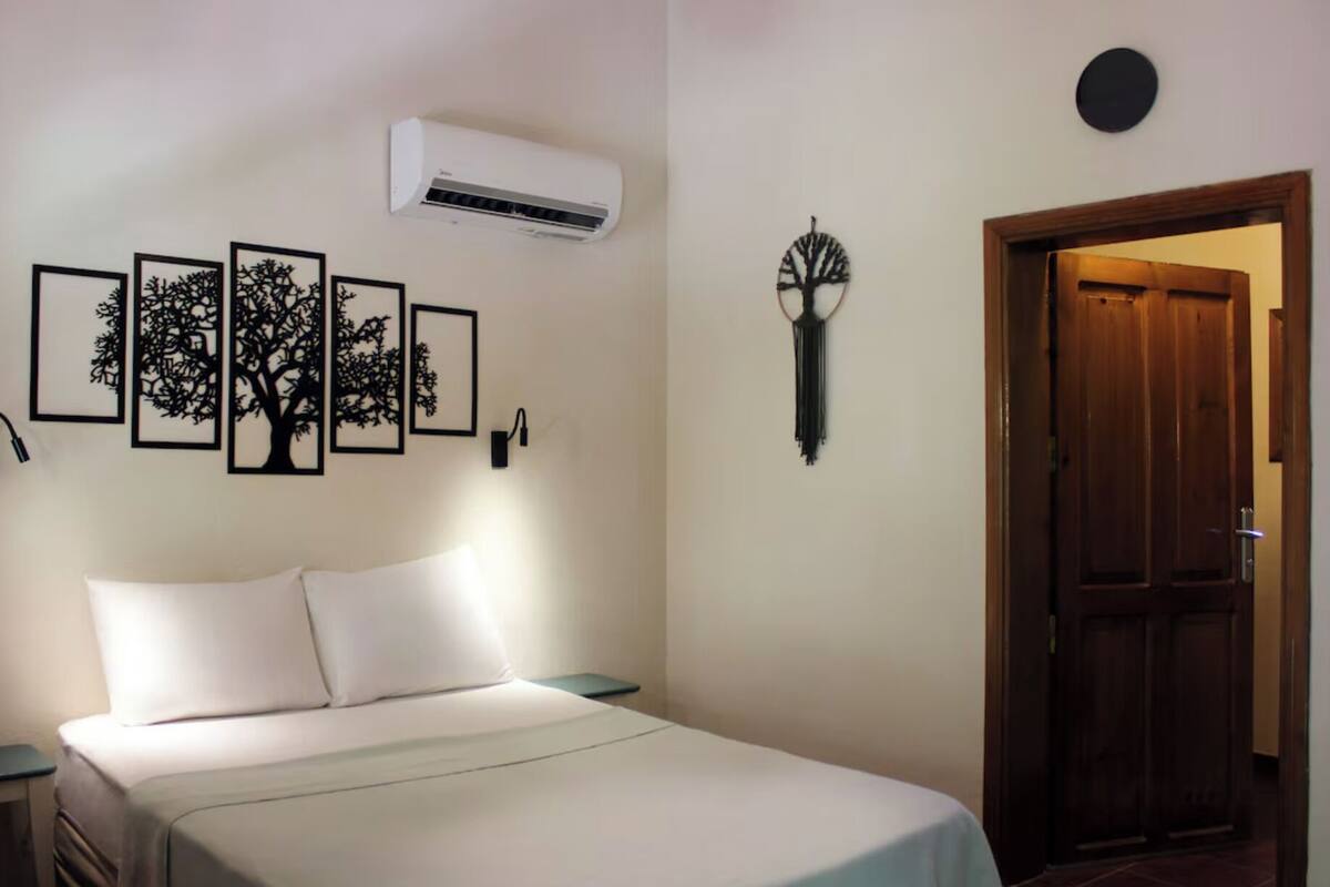 Une chambre à coucher comprenant un lit, un climatiseur fixé au mur, des silhouettes d’arbres encadrées, une lampe fixée au mur et une porte en bois.