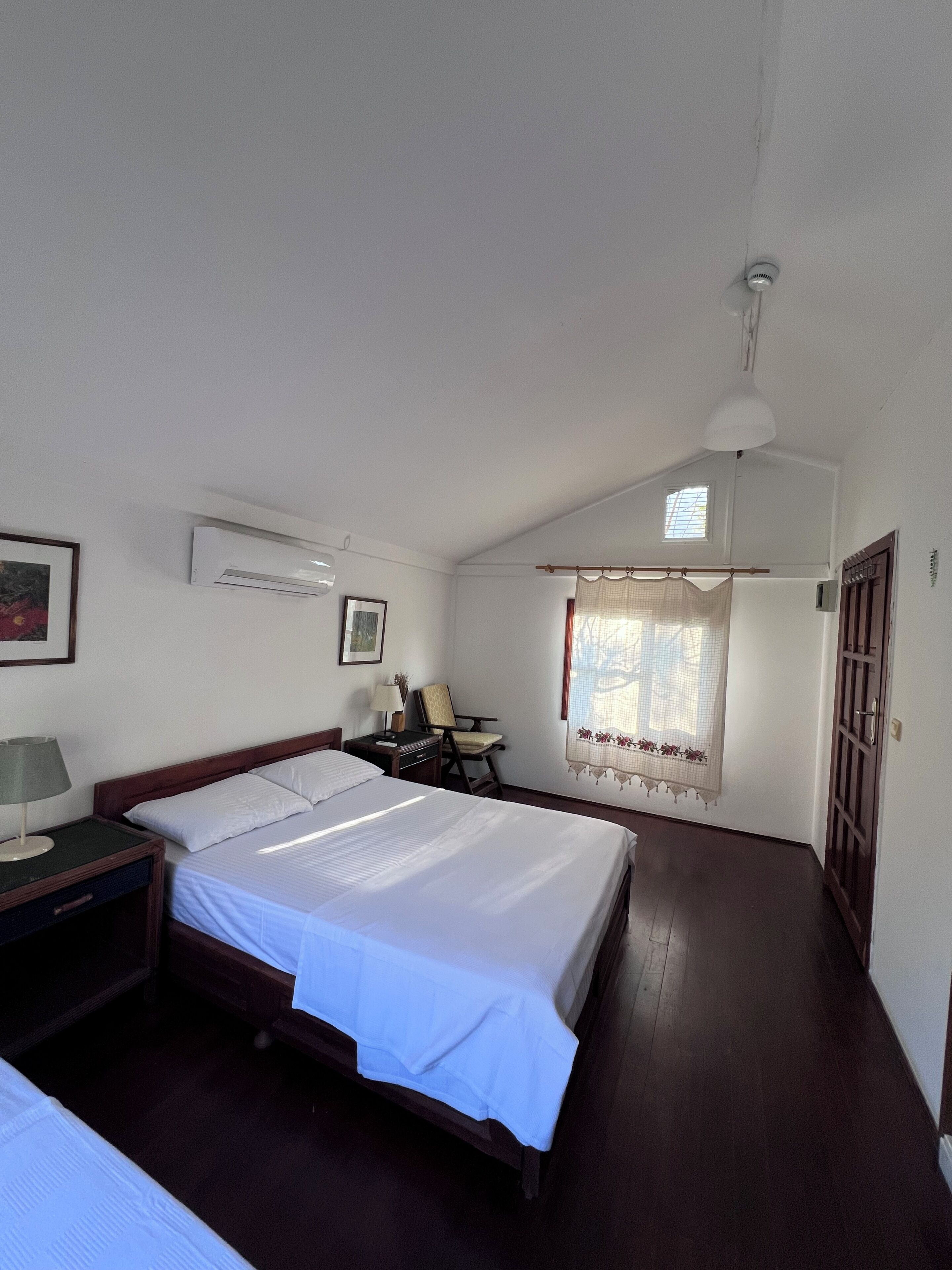 A hotel room with a bed, a desk, a chair, a lamp, a window with curtains, and an air conditioning unit.