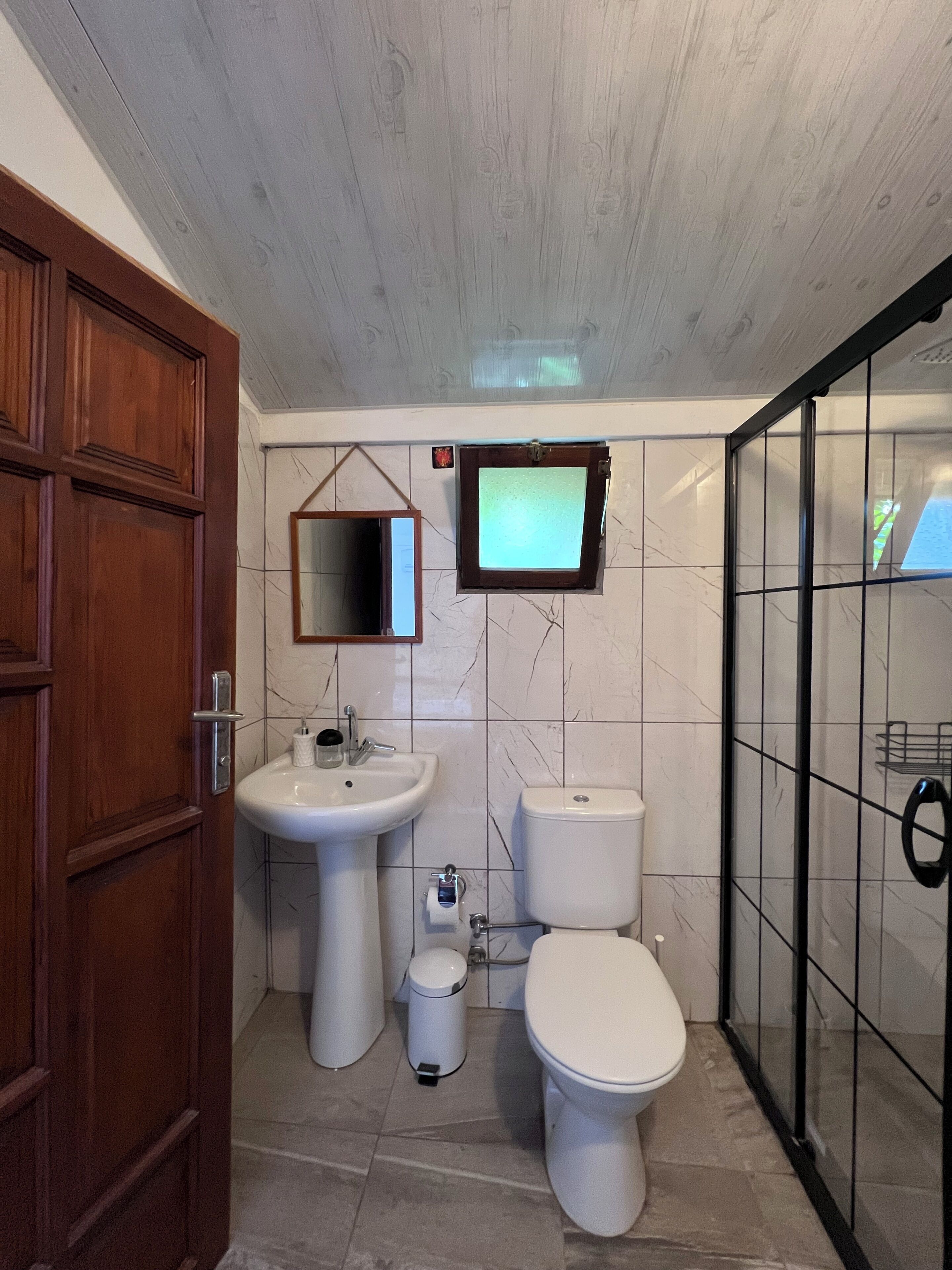 A bathroom with a wooden door, a sink, a toilet, a mirror, and a television mounted on the wall.