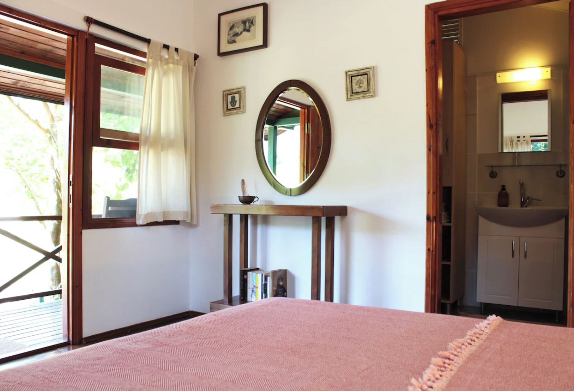 A bedroom with a bed, a bedside table, a mirror, a window with curtains, and a bathroom with a sink and mirror.