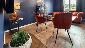 Ein Wohnzimmer mit dunkelblauer Wand, Holzboden, einer Couch, zwei Sesseln, einem Beistelltisch mit Lampe und einem Bücherregal.