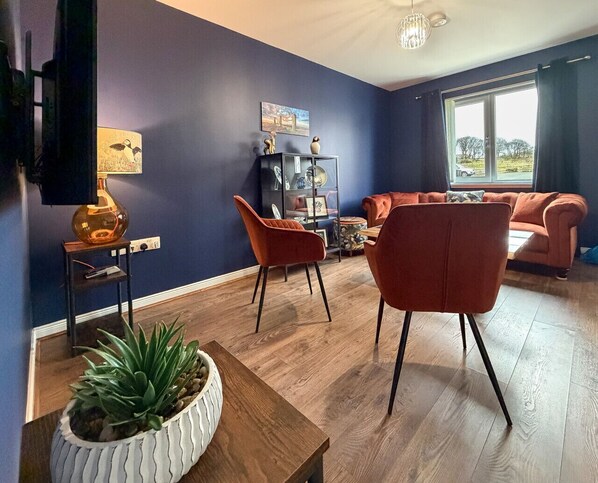 Un salon avec un mur bleu foncé, un plancher en bois, un canapé, deux fauteuils, une table d’appoint avec une lampe et une bibliothèque.