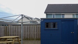 A blue shed, a wooden picnic table, and a metal structure in a backyard with a wooden fence and a house in the background.