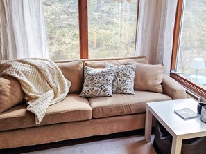 Ein gemütliches Wohnzimmer mit einem beigen Sofa, gestreiften Kissen, einer Decke und einem kleinen Tisch mit Lampe und Buch.