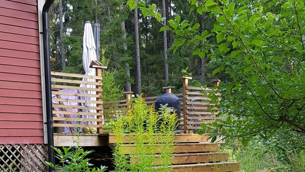 A wooden deck with a grill, surrounded by greenery and a red building. - 4 Star Holiday Home in Mariefred (Mariefred)