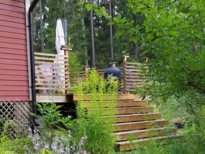Eine Holzterrasse mit Grill, umgeben von Grünpflanzen und einem roten Gebäude.