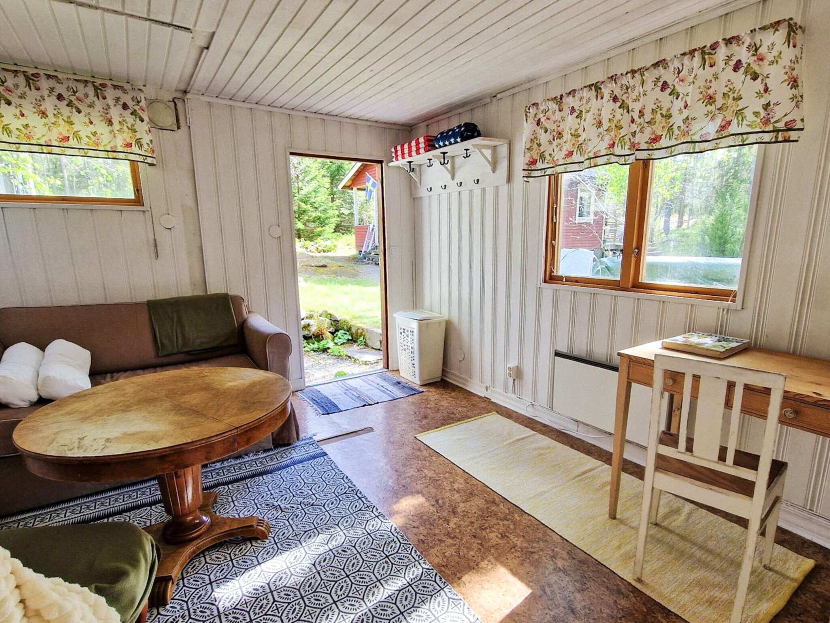 A cozy room with a wooden floor, a brown sofa, a small dining table, and a window with floral curtains.