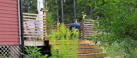 A wooden deck with a grill and a covered area, surrounded by greenery and trees.