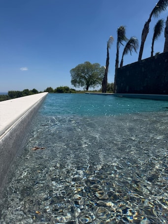 A clear pool with a concrete edge, surrounded by palm trees and a stone wall.. Flora Sicula - Cultivating Hospitality