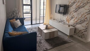 A living room with a blue sofa, a coffee table, a white console, and a TV mounted on a marble-patterned wall. - Crystal Ace Lagoon & Pool Bliss (Johannesburg)