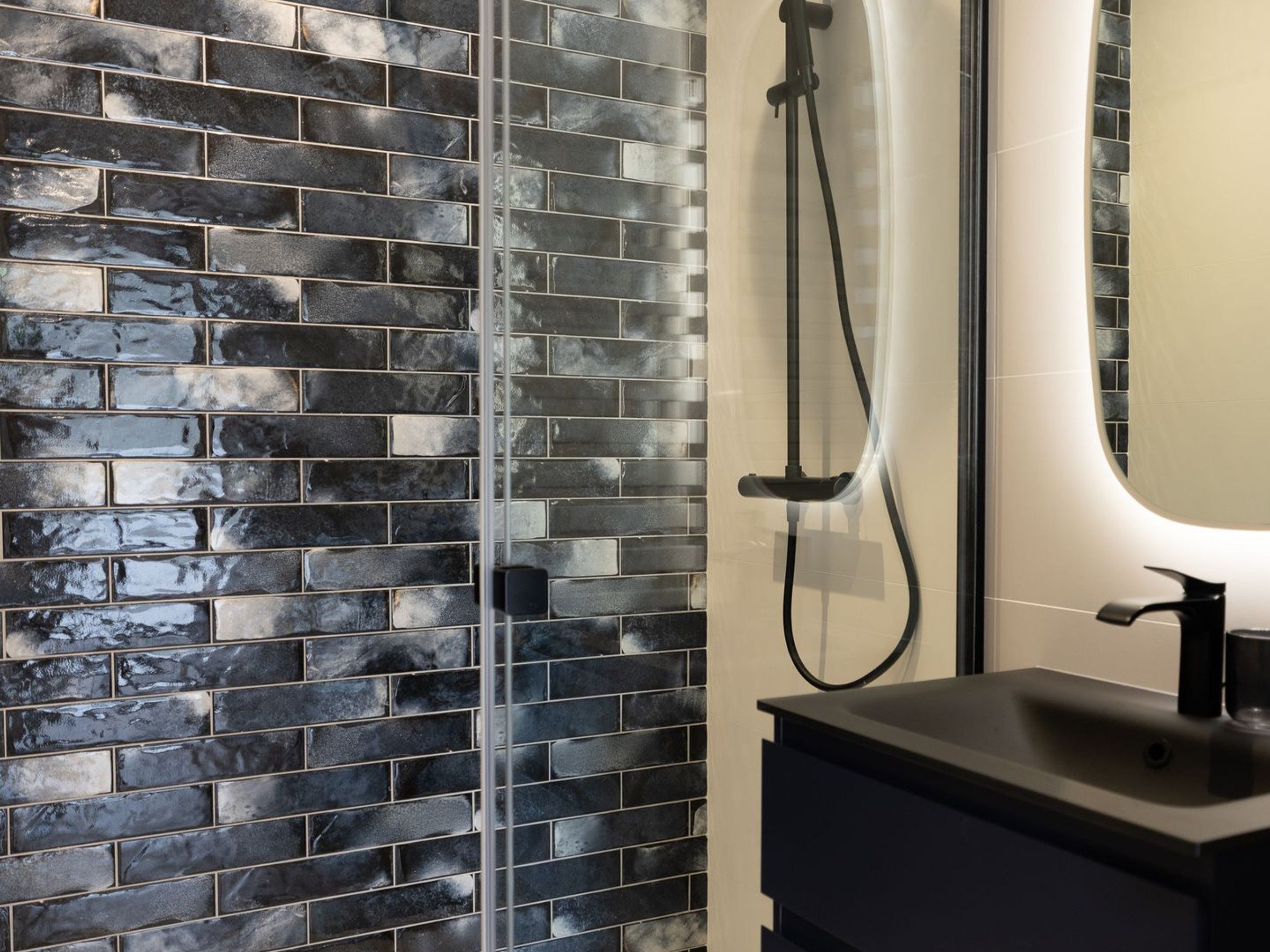 A bathroom with a tiled wall, a shower, and a sink with a mirror.