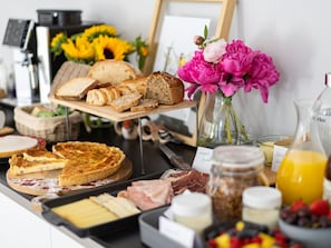 A breakfast buffet with various breads, a quiche, meats, cheeses, and a vase of flowers.